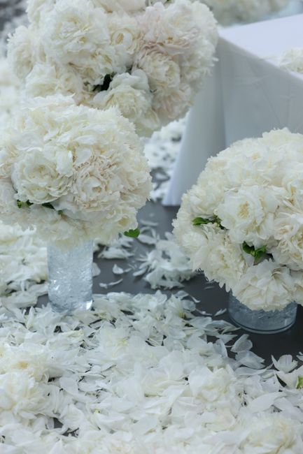 A floral arrangement of large, round white peony bouquets with soft, ruffled petals, placed on a table covered with white flower petals and scattered with peony petals. The bouquets are set in clear g