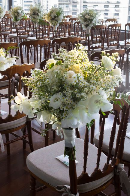 A close-up view of a large, elegant floral bouquet arranged with white roses, orchids, and delicate white filler flowers, featuring lush green foliage. The bouquet is wrapped with a white satin ribbon