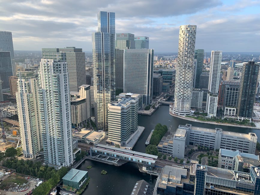 Aerial view of Canary Wharf showcasing a cluster of modern skyscrapers with glass facades reflecting the sky, including a distinctive twisting tower on the right. The buildings surround a waterway wit