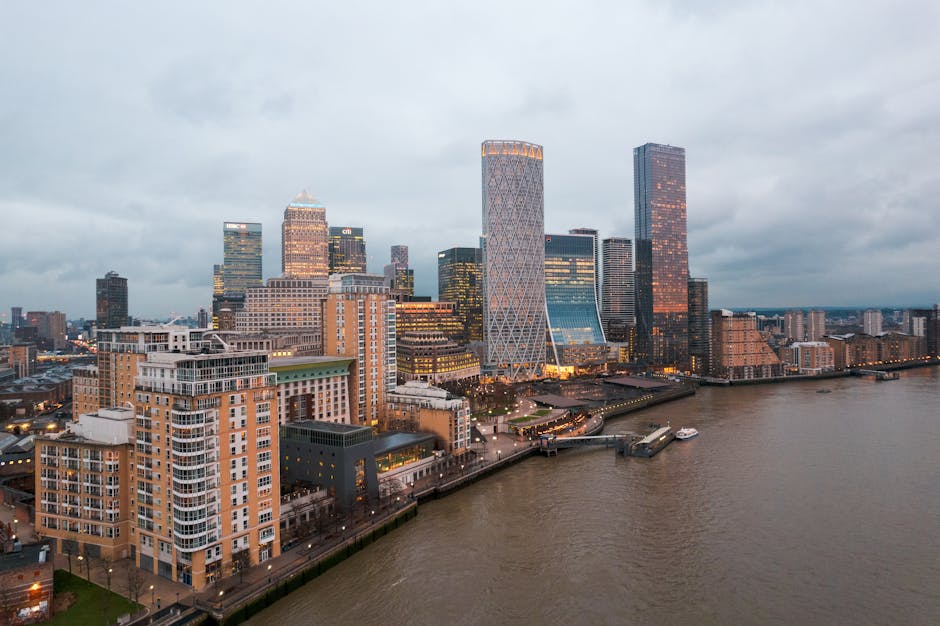 A panoramic view of Canary Wharf's skyline during the evening, featuring modern high-rise office buildings and residential towers with illuminated windows. The waterfront in the foreground shows calm 